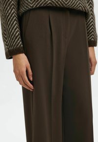 Personne portant un pantalon large marron et un pull marron à motifs, les mains détendues le long du corps, sur un fond clair.