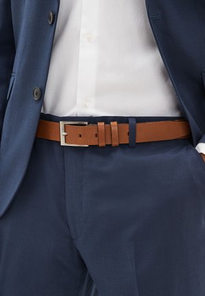 Homme portant un costume bleu marine, une chemise blanche et une ceinture en cuir marron avec une boucle rectangulaire argentée.
