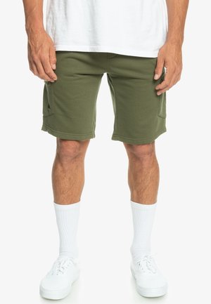 Shorts vert olive avec une coupe décontractée, fabriqués à partir d'un tissu doux, dotés de poches latérales et d'un ourlet élastiqué, associés à des chaussettes blanches et des baskets.