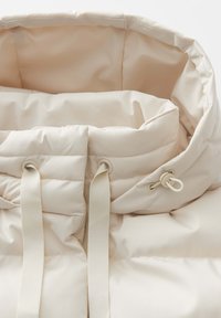 Gepolsterte beige Jackeninnenseite mit hohem Kragen und Kordelzug, glatter Textur und zwei vertikalen Riemen auf der Vorderseite.