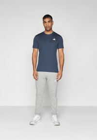 Hombre de pie de frente vistiendo una camiseta azul marina, pantalones de chándal gris claro con franjas laterales y zapatillas deportivas blancas contra un fondo liso.