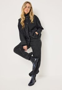 Giacca di jeans nera con pantaloni baggy abbinati, entrambi caratterizzati da una vestibilità ampia. Stivali alla caviglia lucidi di colore blu navy completano l'outfit.