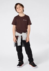 Zēns, kuram mugurā brūna Champion T-kreklu, melni bikses, melnas kedas ar oranžiem akcentiem un pelēks sporta krekls sasietts ap vidu.