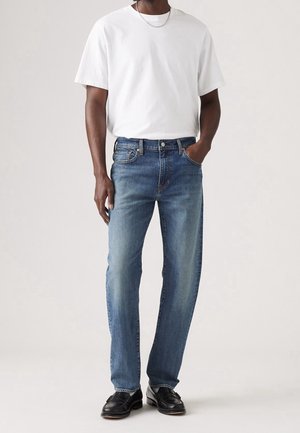 Homme portant un t-shirt blanc à manches courtes, un jean bleu droit, des chaussures en cuir noires et un collier en chaîne argentée, debout avec une main dans la poche.