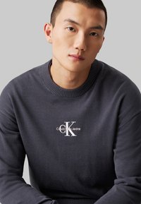 Σκούρο γκρι φούτερ από μαλακό ύφασμα με ριμπ κολάρο. Διαθέτει λευκό ραμμένο λογότυπο "Calvin Klein Jeans" στο στήθος.