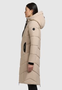 Béžový prešívaný kabát s kapucňou, predstavujúci prešívaný vzor, čierne detaily, bočné vrecká a zips na uzavretie v spodnej časti.