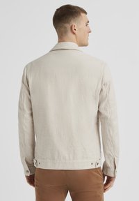 Helle beige Baumwolljacke mit Reverskragen, langen Ärmeln und Knopfdetails an der Taille. Strukturiere Oberfläche mit minimalen Nähten.