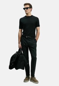 Zwarte ribbelige korte mouwen shirt, zwarte broek, olijfgroene suède schoenen en houdt een zwarte pufferjack vast. Zwarte zonnebril maakt de look compleet.