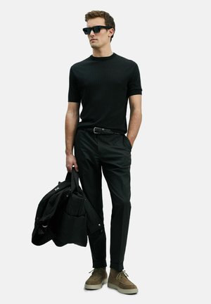 Chemise noire à manches courtes côtelée, pantalon noir, chaussures en suède olive, et tenant un manteau matelassé noir. Des lunettes de soleil noires complètent le look.