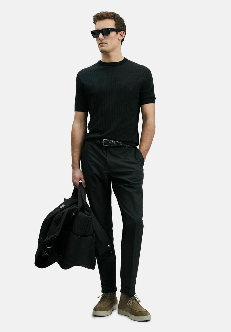 Zwarte ribbelige korte mouwen shirt, zwarte broek, olijfgroene suède schoenen en houdt een zwarte pufferjack vast. Zwarte zonnebril maakt de look compleet.