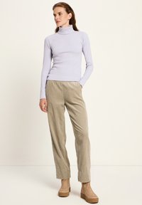 Frau in einem helllila gerippten Rollkragenpullover, beigefarbenen Cordhosen mit Händen in den Taschen und braunen Plateaustiefeln bis zum Knöchel, stehend vor einem schlichten Hintergrund.