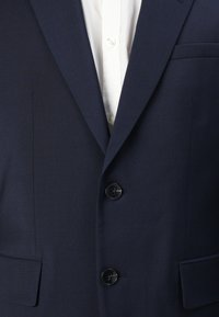 Marineblazer met een gladde textuur, twee zwarte knopen en een enkele borstzak. Wit shirt zichtbaar onder de blazer.
