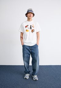 White t-shirt featuring colorful animal and object graphics, paired with light blue jeans and gray-accented sneakers, topped with a plaid bucket hat.