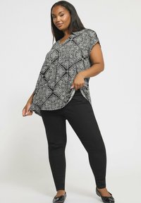 Kvinna klädd i en lös topp med svart och vitt mönster, svarta leggings och svarta ballerinaskor, poserar mot en enfärgad vit bakgrund.