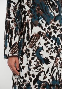 Kootud mantel, millel on julge abstraktne muster teal, pruuni ja valgega. Sellel on mugav lõige, pikad varrukad ja viis musta nööpi.