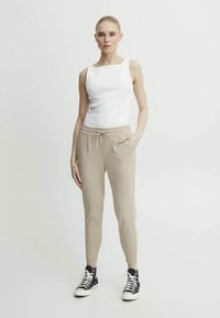 Beige jogger pants with side pockets and a drawstring waist, paired with a fitted white tank top and black high-top sneakers.