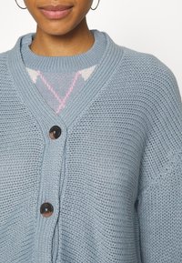 Ljusblå stickad cardigan med V-ringning och ribbad krage, med stora mörka knappar och ett strukturerat mönster. Detaljer av en mönstrad skjorta under.