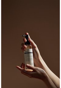 Flaska med Klairs Gentle Black Fresh Cleansing Oil, genomskinlig med en svart pump, innehåller en ljus bärnstenfärgad vätska, hålls i händerna mot en brun bakgrund.