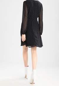 Robe noire à pois blancs avec des manches longues transparentes et une taille cintrée, assortie à des bottines blanches à la cheville, vue de dos.