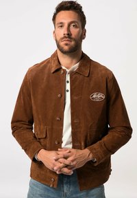 Veste en daim marron avec un col, des boutons à l'avant et deux poches frontales ; présente un logo brodé sur le côté supérieur gauche.