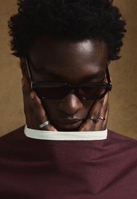 Črna pravokotna sončna očala s temnimi lečami, bordo majica z belim ovratnikom in več kovinskimi prstani na rokah. Vidna je teksturirana koža.