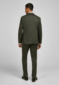 Dunkelgrüner maßgeschneiderter Anzug aus strukturiertem Stoff, mit einem Rückenschlitz, kombiniert mit passenden Hosen und schwarzen Schuhen, von hinten gezeigt.