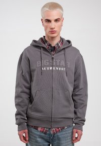 Grå zip hoodie med kangaroo-lommer, med "BIG STAR AUTHENTIC" trykt i lysegrå. Båret over en ternet skjorte, glat tekstur.