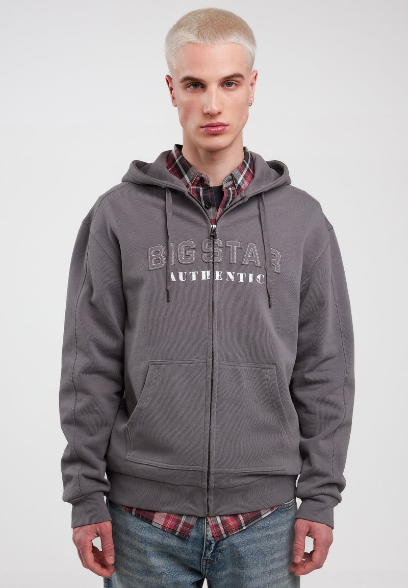 Grå zip hoodie med kangaroo-lommer, med "BIG STAR AUTHENTIC" trykt i lysegrå. Båret over en ternet skjorte, glat tekstur.