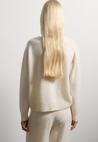 Suéter crema hecho de material suave, con dobladillo y puños acanalados, combinado con pantalones crema a juego. El cabello cae recto por la espalda.