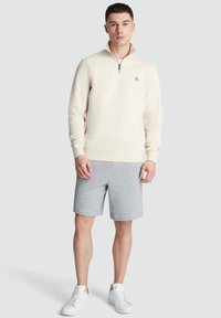 Light beige pullover with a zip collar, soft fabric, ribbed cuffs, and hem. Paired with gray shorts and white sneakers.