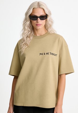 Frau mit langen, welligen blonden Haaren, die schwarze Sonnenbrillen und ein lockeres beiges T-Shirt mit dem Text "YOU & ME TONIGHT" auf der Brust trägt.