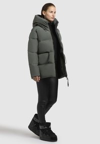 Pufferjack in gedempt groen, met een capuchon en ritssluiting, gecombineerd met zwarte leren leggings en zwarte hoge sneakers.