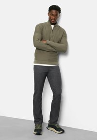 Olivengrüner Zip-Hals-Pullover, strukturiertes Material, über einem weißen Hemd getragen, kombiniert mit tiefschwarzen Hosen und dunklen Sneakers mit Akzenten.