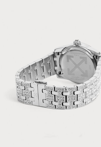 Montre-bracelet en métal argenté avec un bracelet à maillons comportant des strass clairs incrustés et un boîtier arrière rond avec des flèches gravées.