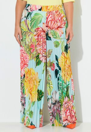 Kolorowe plisowane spodnie o szerokich nogawkach z dużym kwiatowym wzorem w żółtym, różowym i zielonym kolorze, noszone z żółtą bluzką i pomarańczowymi, spiczastymi butami.