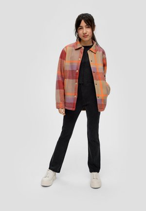 Plaid shirt jacket in shades of orange, pink, and beige, with a collared neckline and snap buttons. Paired with black textured pants and white sneakers.