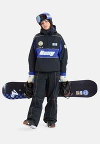 Snowboardzista nosi czarną i niebieską kurtkę z zamkiem błyskawicznym, z naszywkami i kapturem, czarne spodnie i rękawice, trzymając snowboard z wiązaniami.