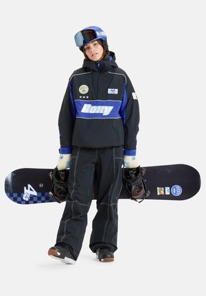 Snowboardzista nosi czarną i niebieską kurtkę z zamkiem błyskawicznym, z naszywkami i kapturem, czarne spodnie i rękawice, trzymając snowboard z wiązaniami.