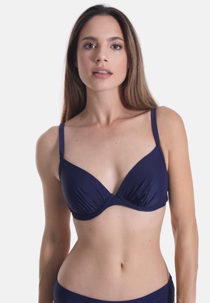 Donna con capelli castani lunghi che indossa un top bikini blu scuro, in piedi su uno sfondo bianco uniforme.