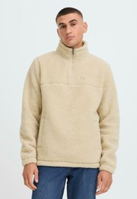 Beige fleece pullover med halv-zip krave, tekstureret stof og en vandret søm over brystet. Har en frontlomme og ribbede manchetter.