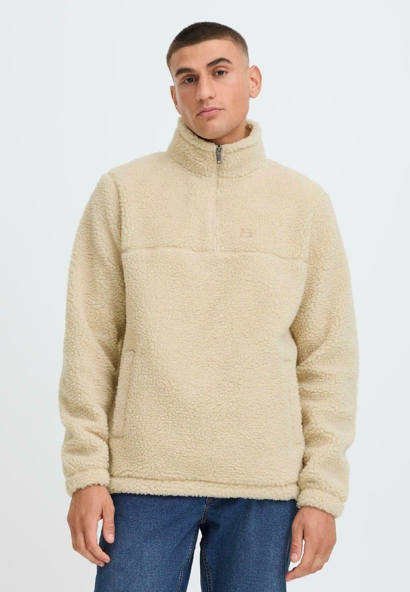 Beige fleece pullover med halv-zip krave, tekstureret stof og en vandret søm over brystet. Har en frontlomme og ribbede manchetter.