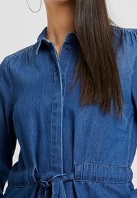Abito in denim blu scuro con colletto, chiusura con bottoni, spalle arricciate e cintura in vita per una vestibilità regolabile.