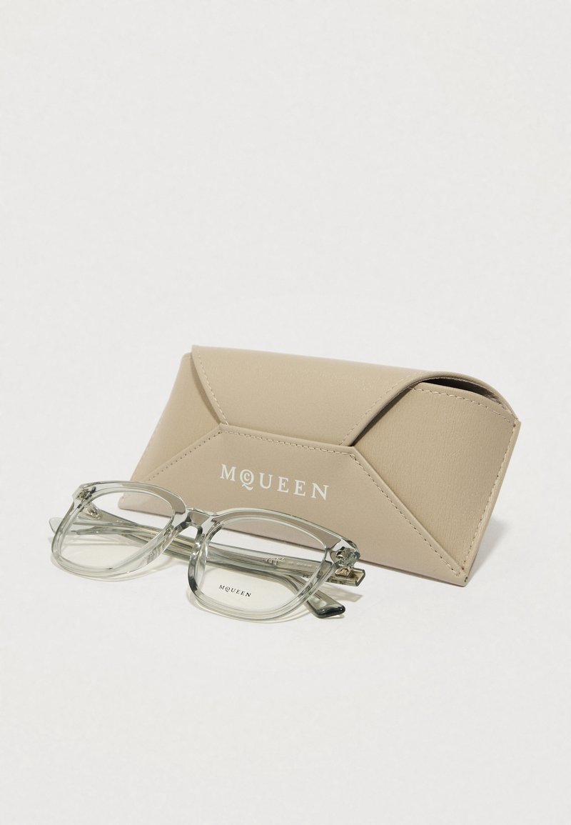 Gafas rectangulares transparentes con un marco ligero reposando sobre un estuche texturizado beige que muestra "MQUEEN" en blanco.