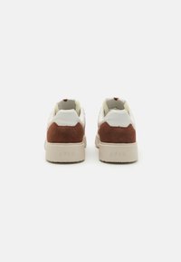Weiße und braune Wildledersneaker mit einem strukturierten Design, gepolstertem Kragen und einer beigen Gummisohle. ARKK-Logo auf der Ferse sichtbar.