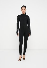 Svart helkropps jumpsuit med lange blonder ermer, høy krage, glidel�ås lukking og tettsittende design, med en subtil preget logo på brystet.
