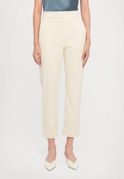 Woman wearing high-waisted, cream-colored straight-leg cropped trousers with side pockets and white pointed-toe slip-on shoes.