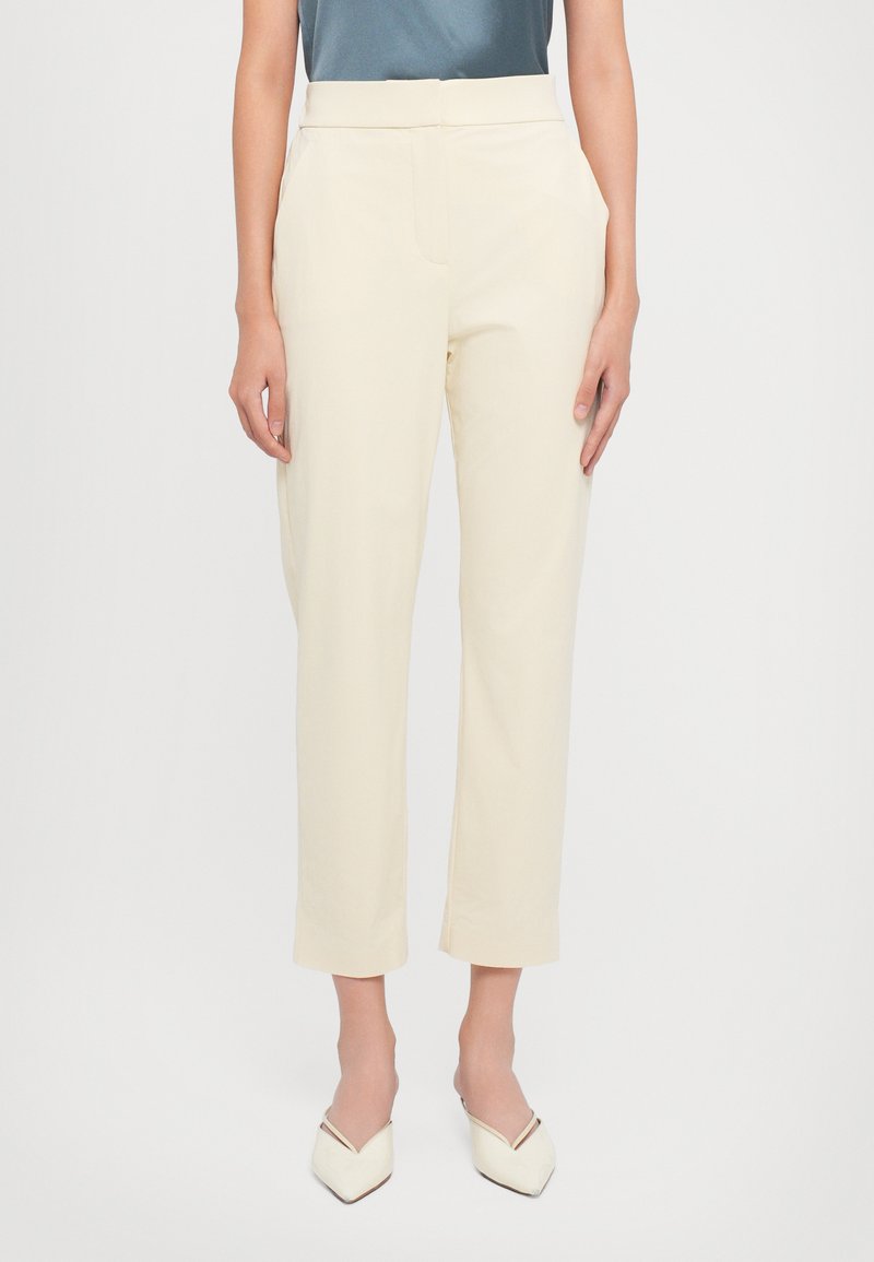 Mulher vestindo calças curtas de perna reta de cintura alta, na cor creme, com bolsos laterais e sapatos de apontado branco e estilo slip-on.