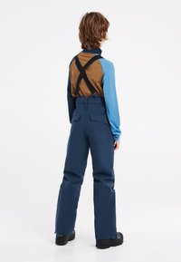 Blauwe langemouwen shirt met bruine accenten, gedragen met marineblauwe tuinbroek. De tuinbroek heeft verstelbare zwarte banden en brede broekspijpen.