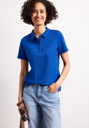 Lachende vrouw met krullend haar, gekleed in een blauwe polo en lichtblauwe jeans, houdt een zonnebril en een bruine armband om haar pols.