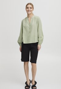 Hellgrünes Leinenblusenshirt mit V-Ausschnitt, Knopfleiste und gerafften langen Ärmeln, kombiniert mit schwarzen knielangen Shorts und flachen Sandalen.